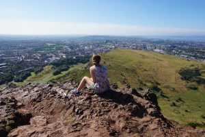 A girl looking over Edinburgh, Scotland while sitting on the top of a mountain in the sun.