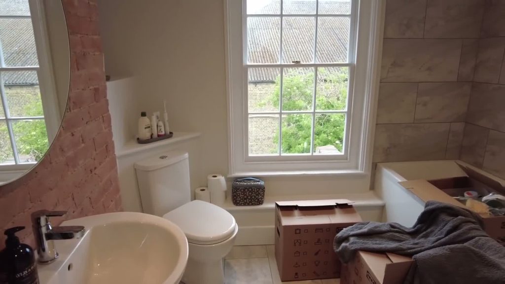 A toilet beside a low uncovered bathroom window, with moving boxes in the room.