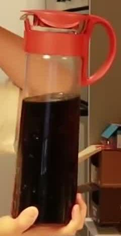 Tall glass jug partly filled with cold brew coffee, with a red lid and handle