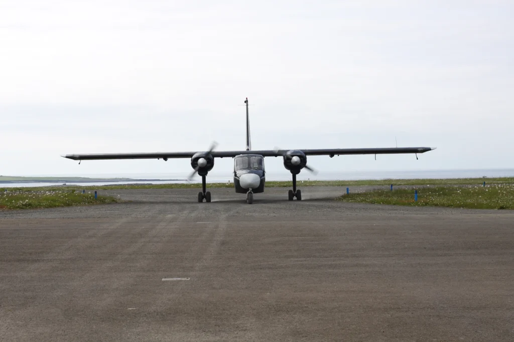 A twin-engine Loganair Islander facing the camera at Papa Westray, with the sea behind.