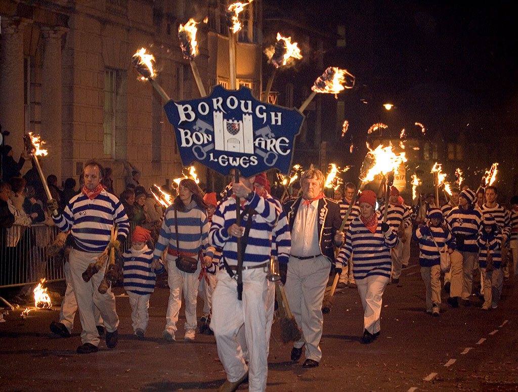 Lewes Borough Bonfire Society members in blue and white striped clothing marching behind a banner and carrying flaming torches.
