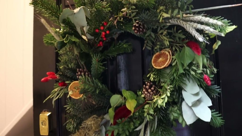 A homemade Christmas wreath with greenery, pine cones and dried orange slices hanging on a dark door.