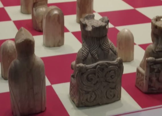 Backs of carved ivory Lewis Chessmen, including patterned chairs, on a red-and-white board.