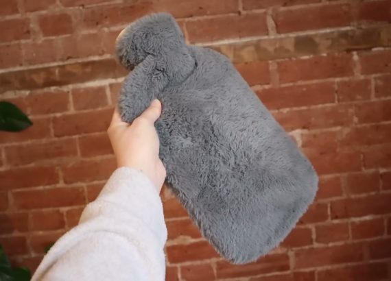A hand holds a hot water bottle in a grey furry cover against a brick wall.