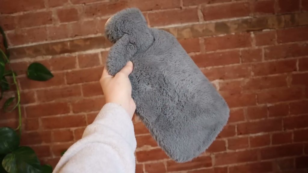 A hand holds a hot water bottle in a grey furry cover against a brick wall.