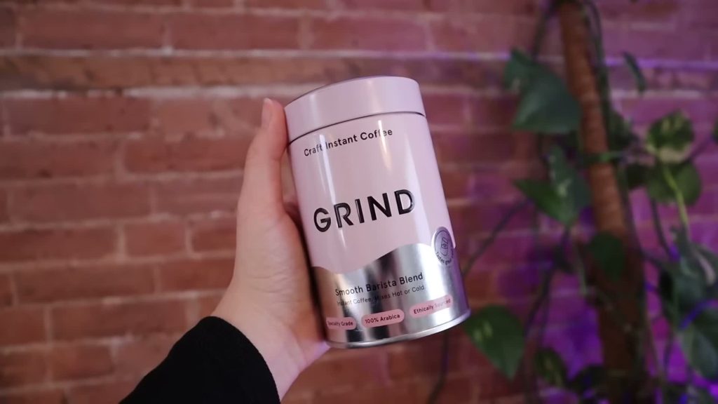 A hand holds the pink and silver Grind Smooth Barista Blend instant-coffee tin.