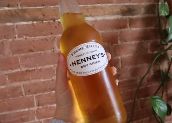 A bottle of Henney's dry cider held in front of a brick wall.