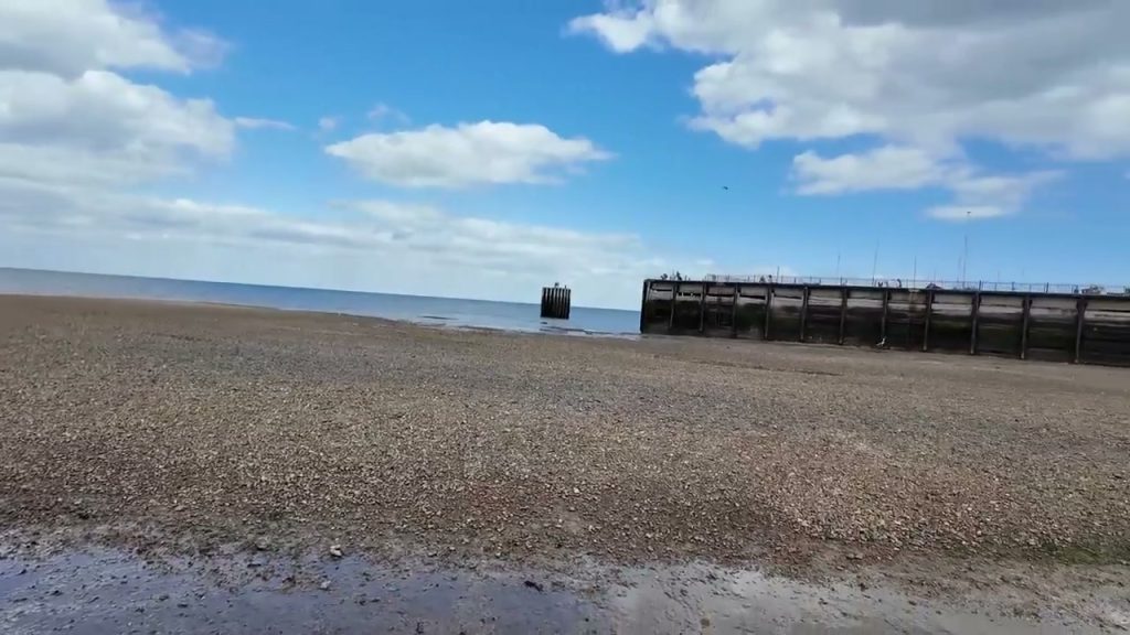 A broad shingle beach and harbour structure at Whitstable under a cloudy sky.