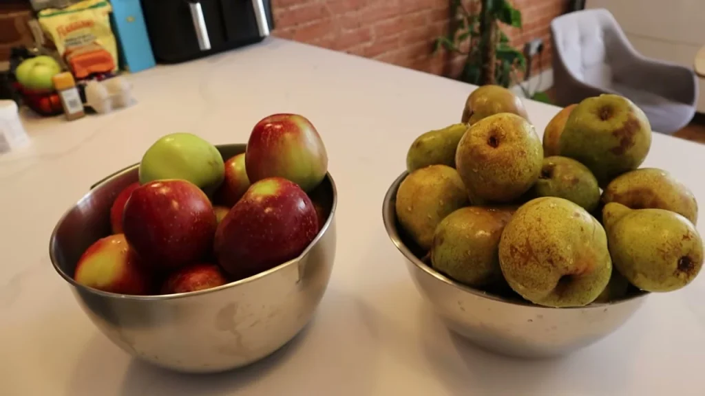 Two metal bowls on a kitchen counter, one filled with apples and the other with pears.